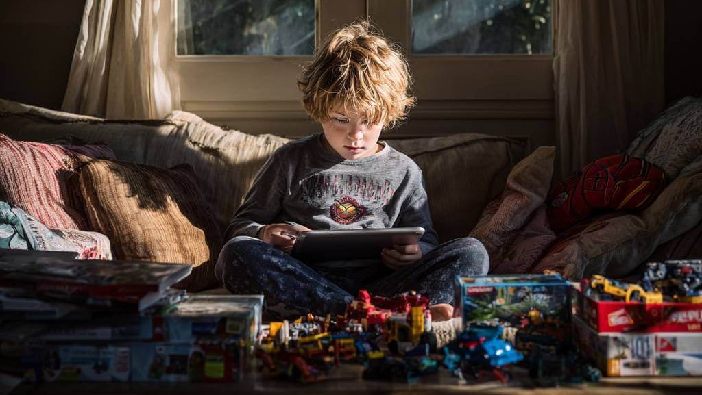 Niño sentado en el sofá usando una tablet, rodeado de juguetes sin tocar en una sala iluminada por la luz natural.