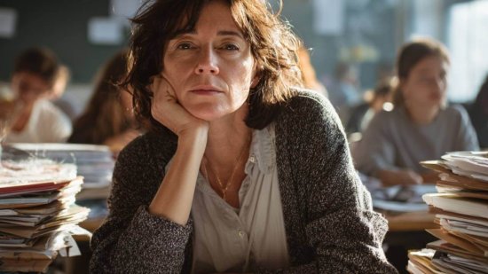 Profesora cansada en el aula, con expresión de agotamiento y pilas de documentos sobre la mesa.