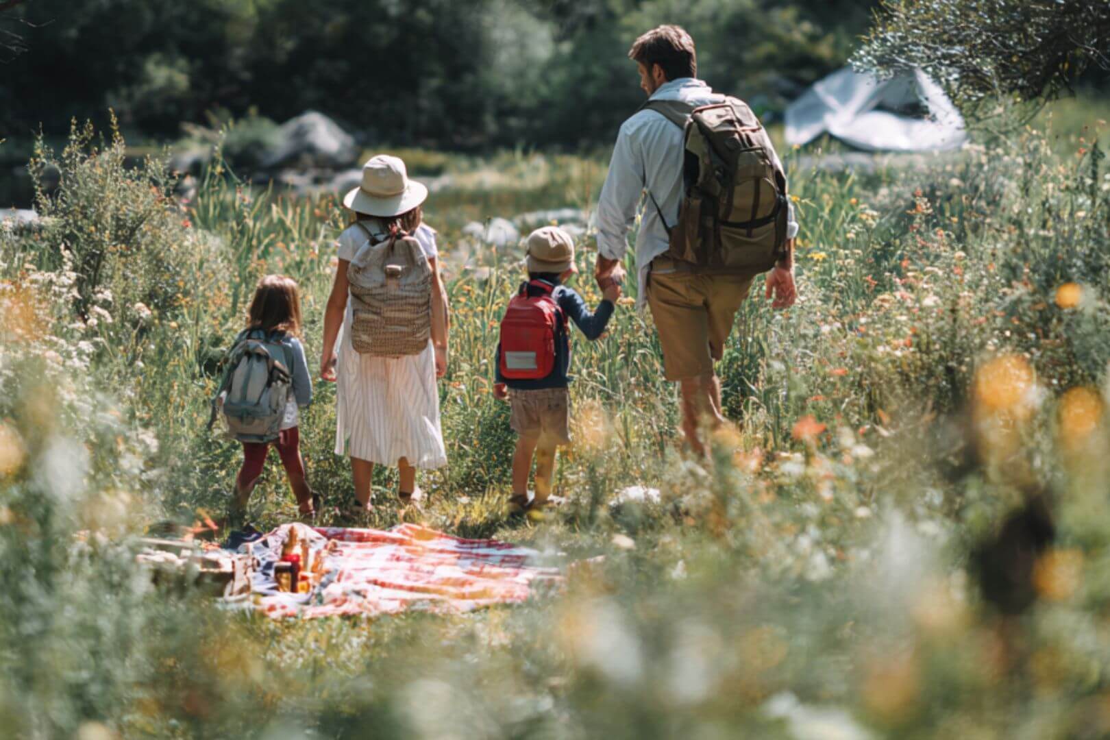 Padres e hijos caminan juntos por un sendero rodeado de flores y hierba alta, llevando mochilas y explorando la naturaleza en familia.