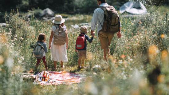 Padres e hijos caminan juntos por un sendero rodeado de flores y hierba alta, llevando mochilas y explorando la naturaleza en familia.