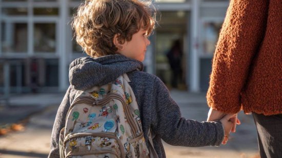 Niño con mochila tomada de la mano de un adulto en la entrada del colegio, símbolo de acompañamiento en la vuelta al cole.