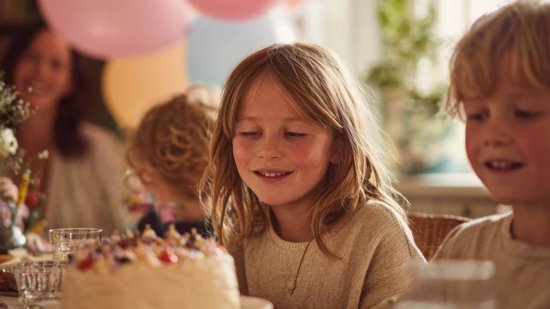 Niños sonríen alrededor de una mesa en una fiesta de cumpleaños, con una tarta decorada y globos al fondo en un ambiente festivo y hogareño.