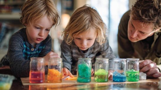 Dos niños pequeños observan frascos con líquidos de colores junto a un adulto, realizando un experimento casero STEAM en un entorno familiar.