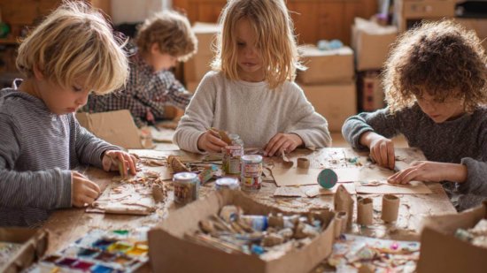 Grupo de niños sentados alrededor de una mesa haciendo manualidades con cartón, pinturas y materiales reciclados en un entorno hogareño y creativo.