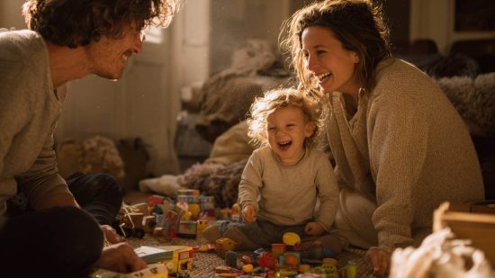 Padres jugando y riendo con su hijo de dos años en casa, rodeados de juguetes y luz cálida de tarde.