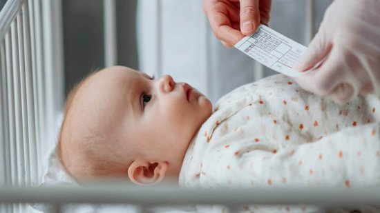 personal sanitario sostiene una tarjeta de cribado genético junto a un recién nacido en una cuna hospitalaria.