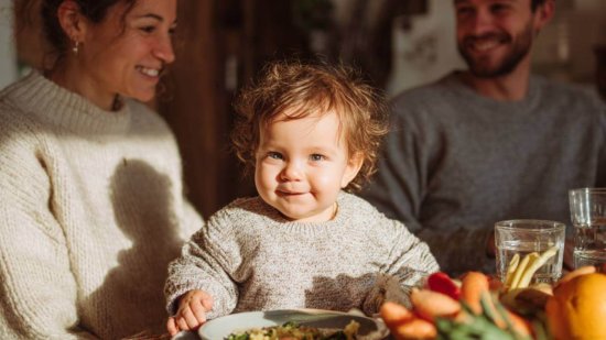 Bebé sonriente sentado a la mesa con sus padres, rodeado de frutas y verduras frescas durante la comida familiar.