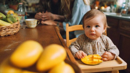 Bebé sentado en su trona probando por primera vez trocitos de mango maduro durante la alimentación complementaria con método BLW, en una cocina familiar y acogedora.