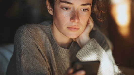 Mujer joven con gesto preocupado mirando su teléfono móvil en un ambiente cálido y con luz suave.