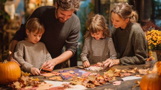 Familia con niños haciendo manualidades de Halloween en casa con calabazas y hojas otoñales.